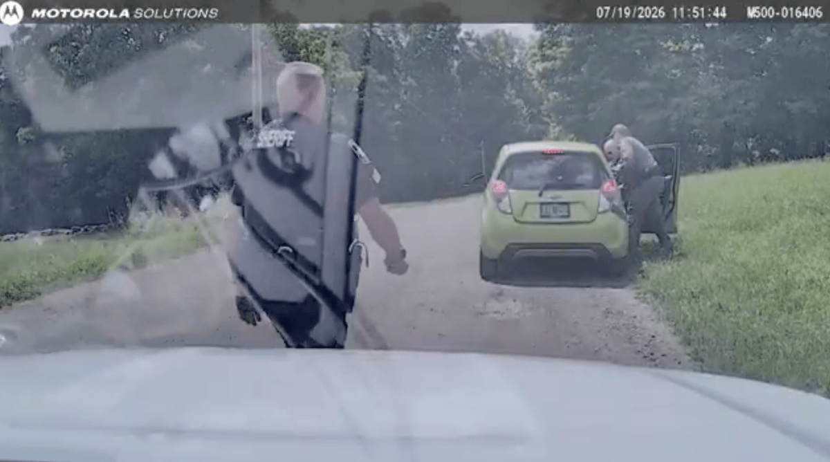 Two police officers approach a green car stopped on a rural dirt road in daylight; one officer stands by the driver’s door, another walks from a patrol vehicle.