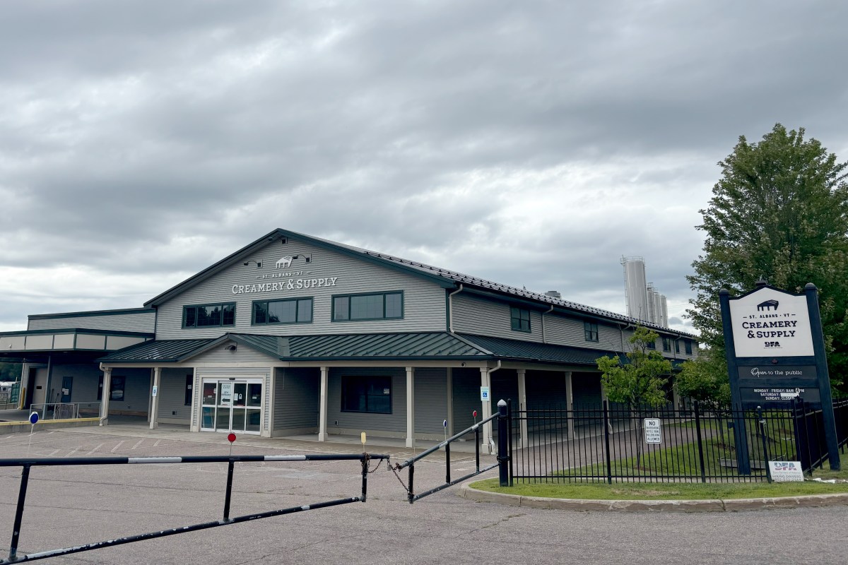 Gray building labeled “Creamery & Supply” sits behind a gated entrance, with a sign near the street. Overcast sky above and trees to the right.