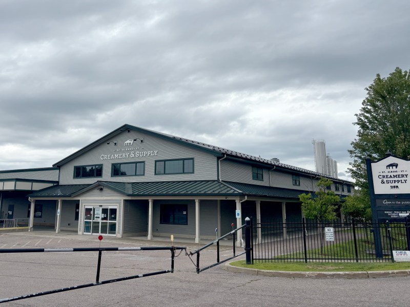 Gray building labeled “Creamery & Supply” sits behind a gated entrance, with a sign near the street. Overcast sky above and trees to the right.