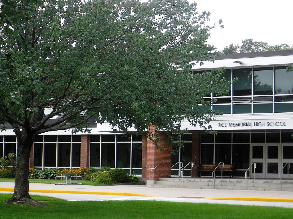A modern, two-story school building with large windows and a sign reading “Rice Memorial High School.” A tree and bike racks are visible in front.