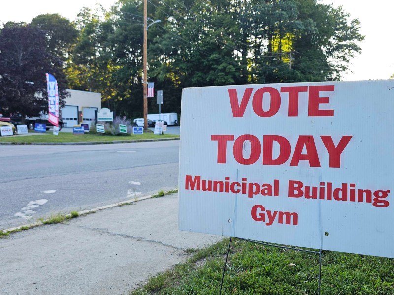 Large sign reads “VOTE TODAY Municipal Building Gym” near a road lined with election campaign signs and trees in the background.
