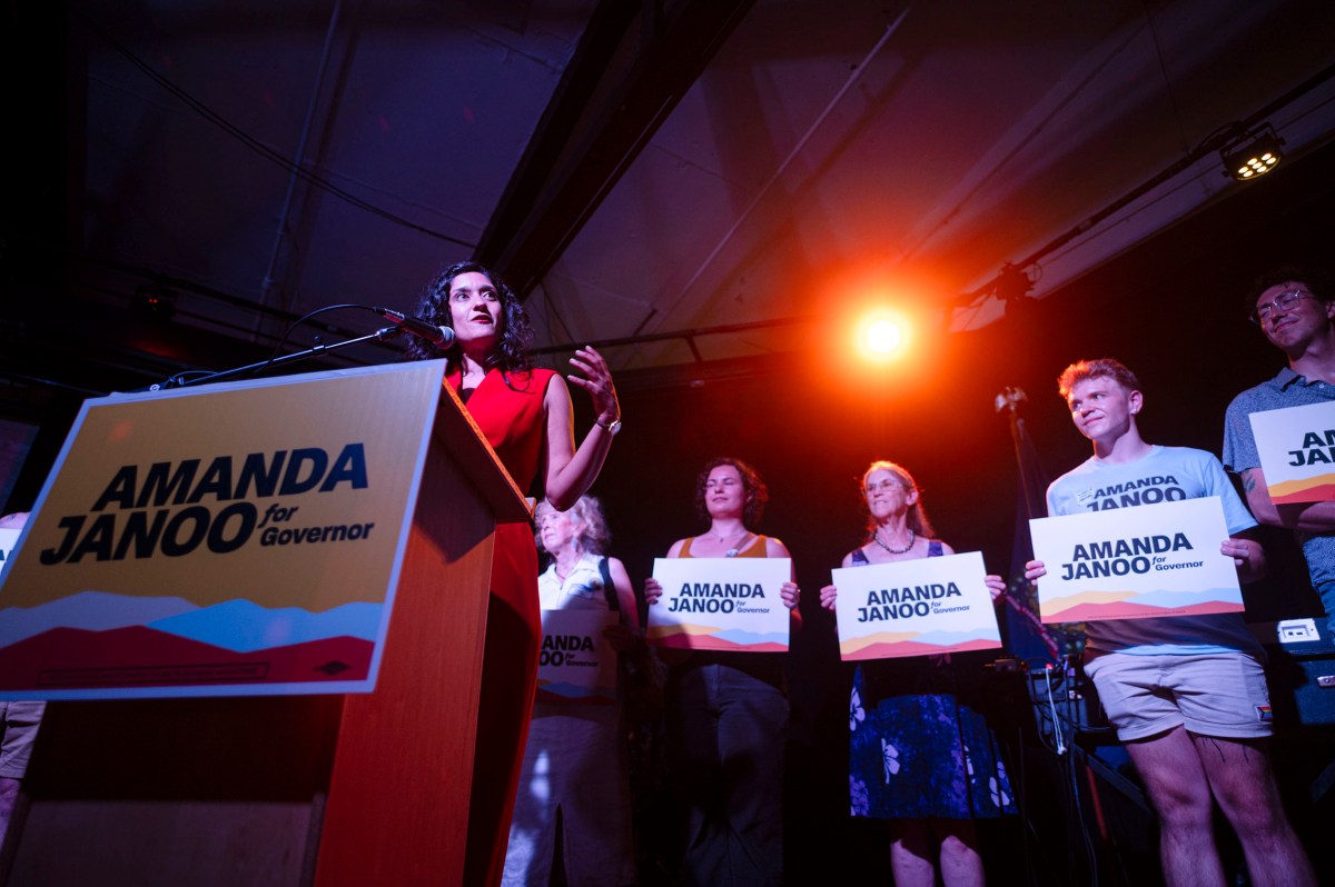 A woman speaks at a podium labeled “Amanda Janoo for Governor” while several people stand beside her holding campaign signs.