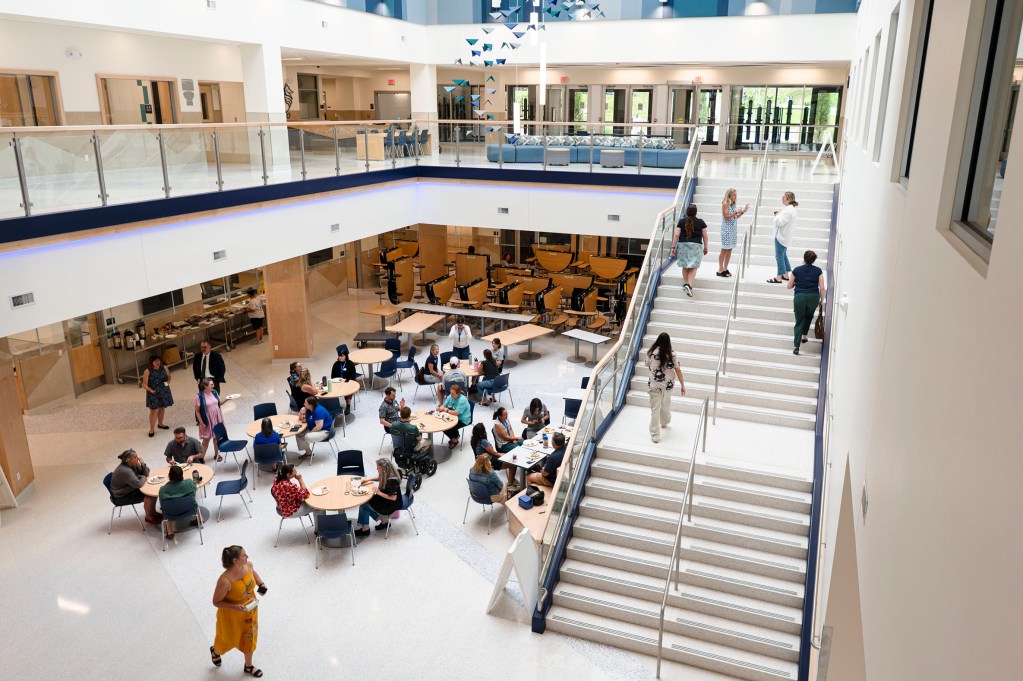 People are gathered at tables and walking in a spacious, modern atrium with large stairs and glass walls in a public building.