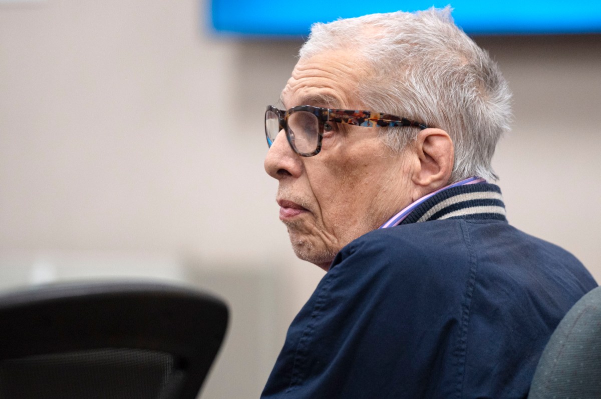 An older man with gray hair and glasses wearing a blue jacket sits at a table, looking to the side in an indoor setting.