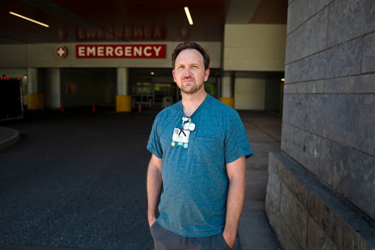 A person wearing a blue shirt and hospital ID stands outside near an emergency entrance, looking at the camera.