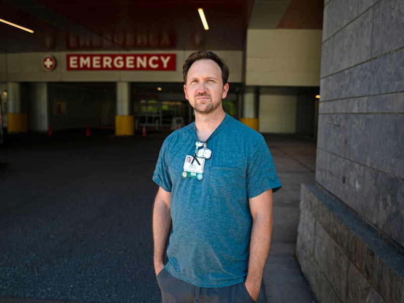 A person wearing a blue shirt and hospital ID stands outside near an emergency entrance, looking at the camera.