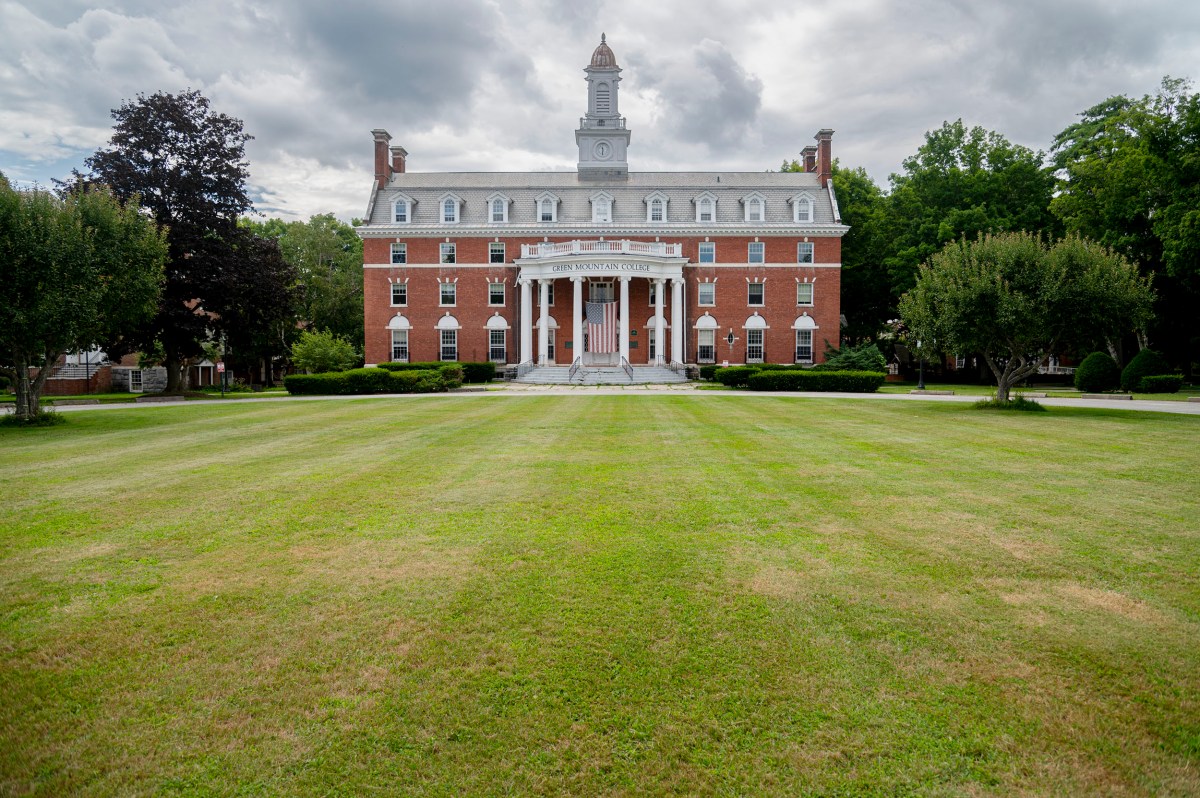 A large brick academic building with white columns stands behind a wide, neatly mowed lawn, surrounded by trees under a cloudy sky.