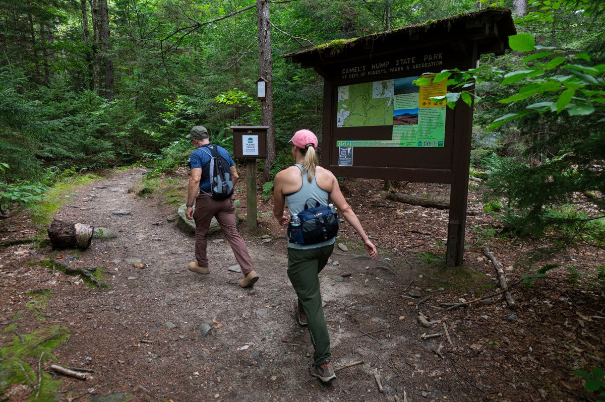 Two hikers with backpacks walk past a trailhead sign and map in a wooded area at Camel’s Hump State Park.