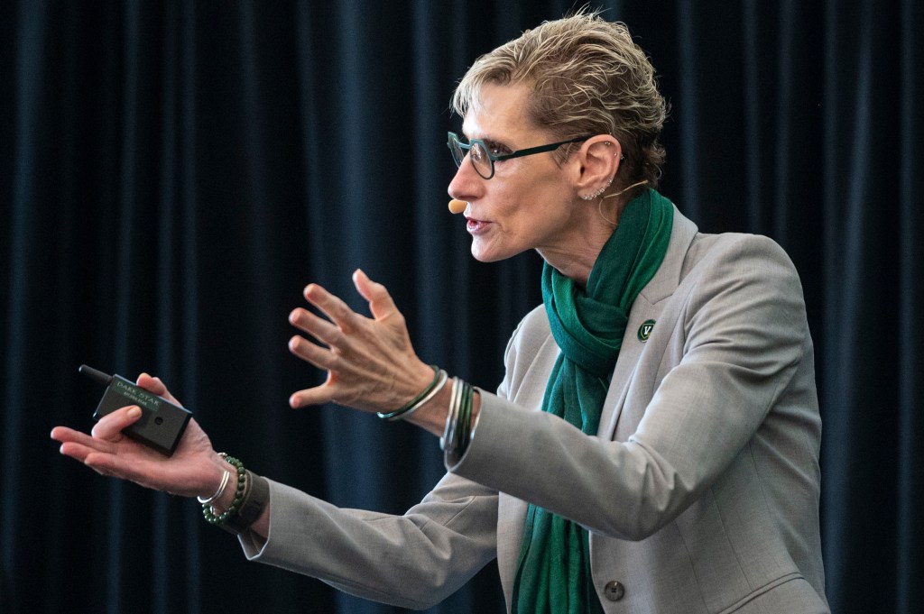 Person wearing glasses and a headset microphone gestures while speaking, dressed in a light blazer and green scarf, standing in front of a dark curtain.