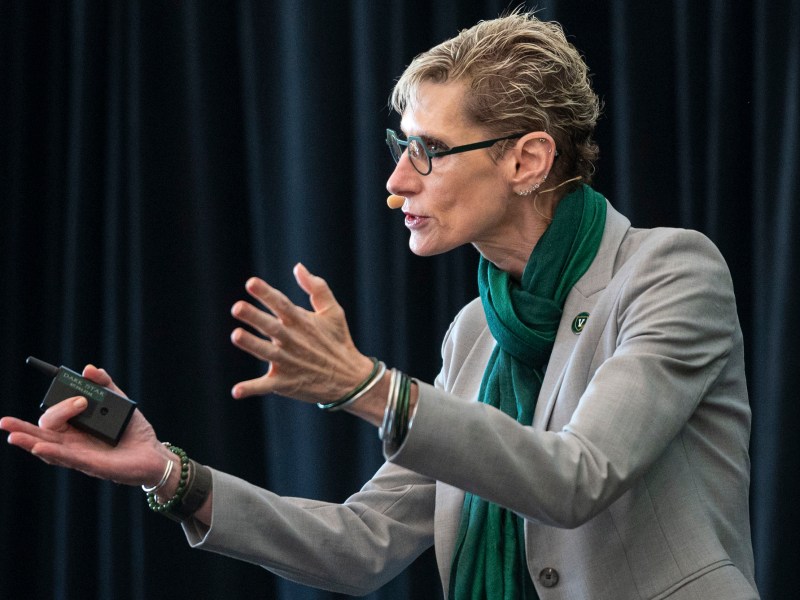 Person wearing glasses and a headset microphone gestures while speaking, dressed in a light blazer and green scarf, standing in front of a dark curtain.
