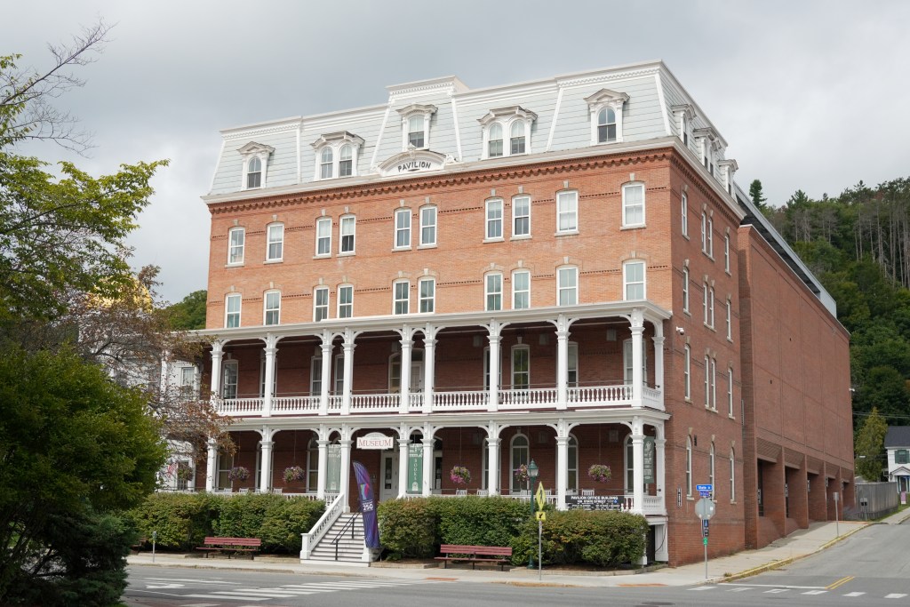 A large, four-story brick building with white-trimmed windows and a covered porch, housing a museum, stands on a corner with trees and benches nearby.