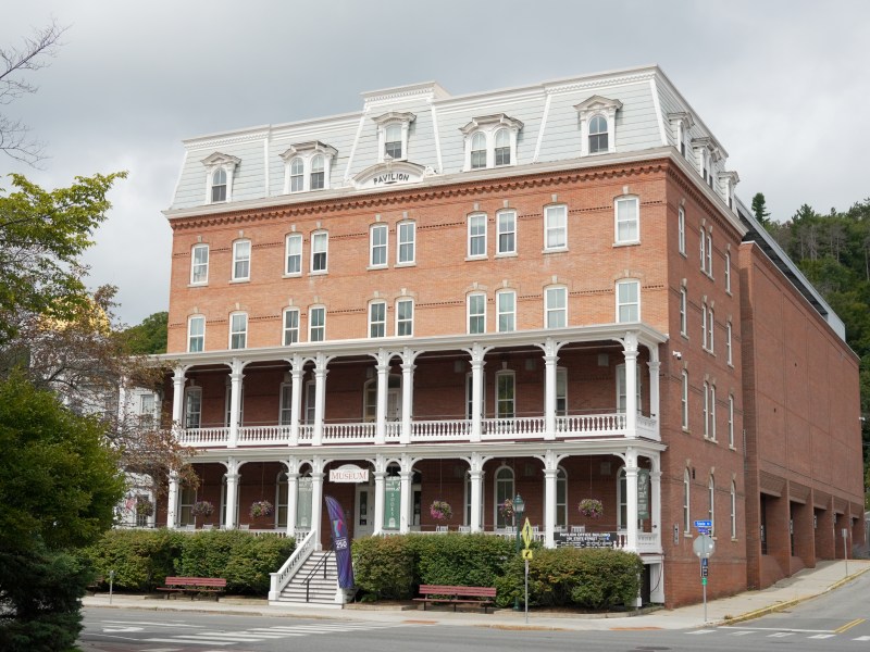 A large, four-story brick building with white-trimmed windows and a covered porch, housing a museum, stands on a corner with trees and benches nearby.