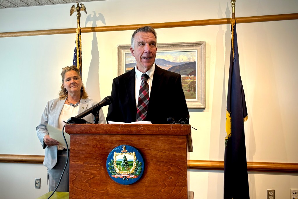 A man in a suit speaks at a podium with a seal, while a woman stands nearby holding papers. Flags and a framed painting are in the background.