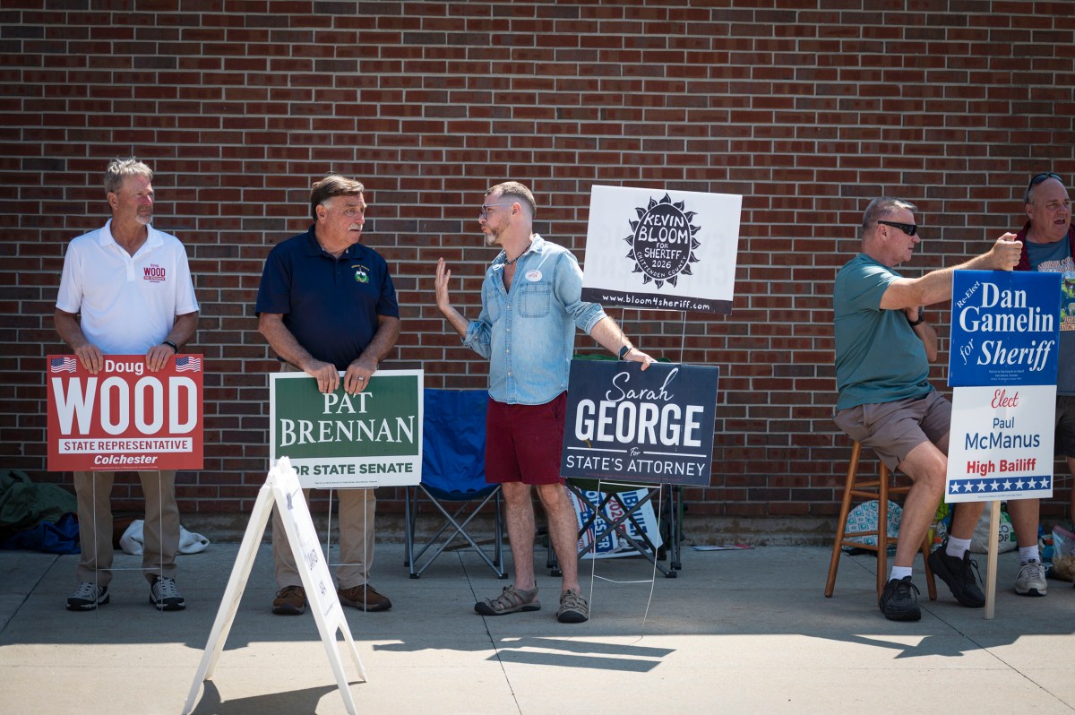 Five men stand on a sidewalk holding signs for various political candidates in front of a brick wall on a sunny day.