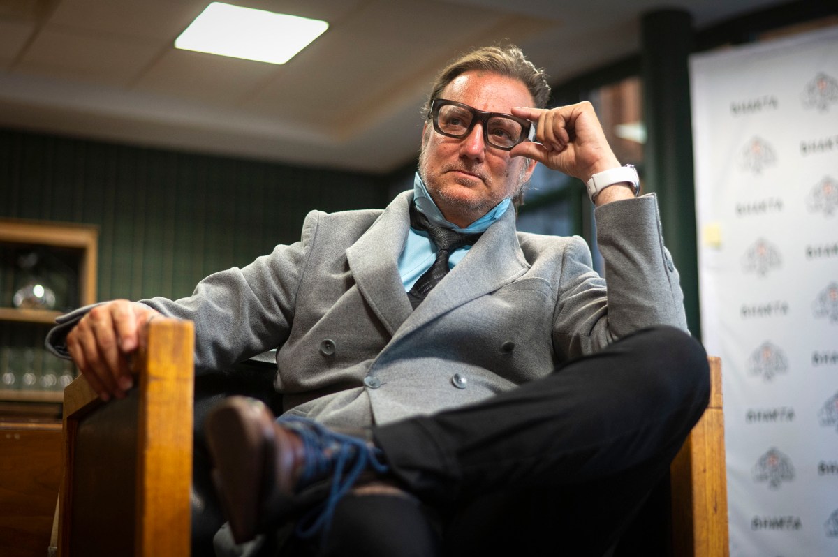 A man in a gray coat and glasses sits in a wooden chair with his legs crossed, resting his hand on his face in a thoughtful pose.