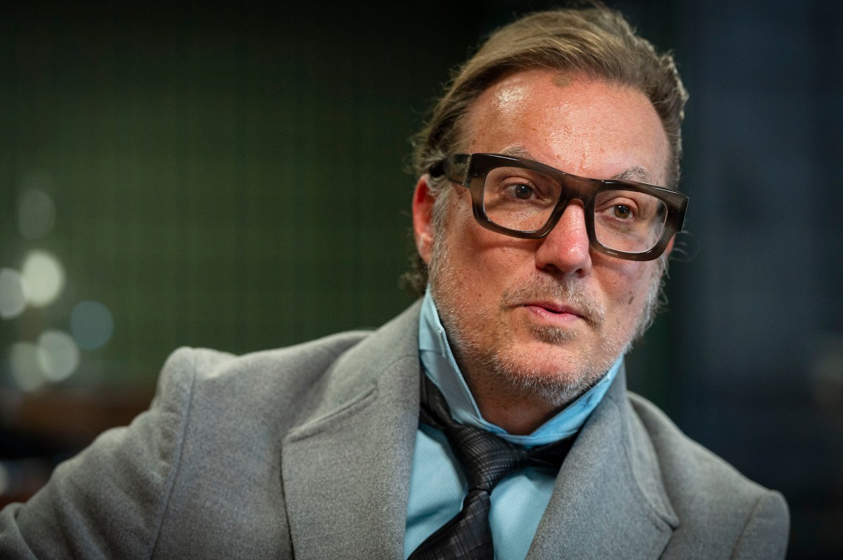 A man in a gray suit and black tie wears large glasses sitting indoors with a blurred green background.