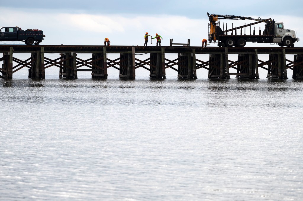 Four workers in safety gear work on a wooden bridge over water, with two trucks parked at opposite ends of the bridge under a cloudy sky.