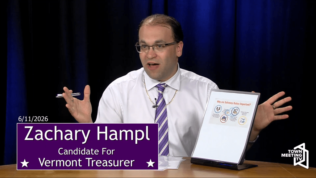 A man in a white shirt and striped tie speaks while seated at a desk with a sign reading “Zachary Hampl, Candidate for Vermont Treasurer.” He gestures toward a chart on a display.
