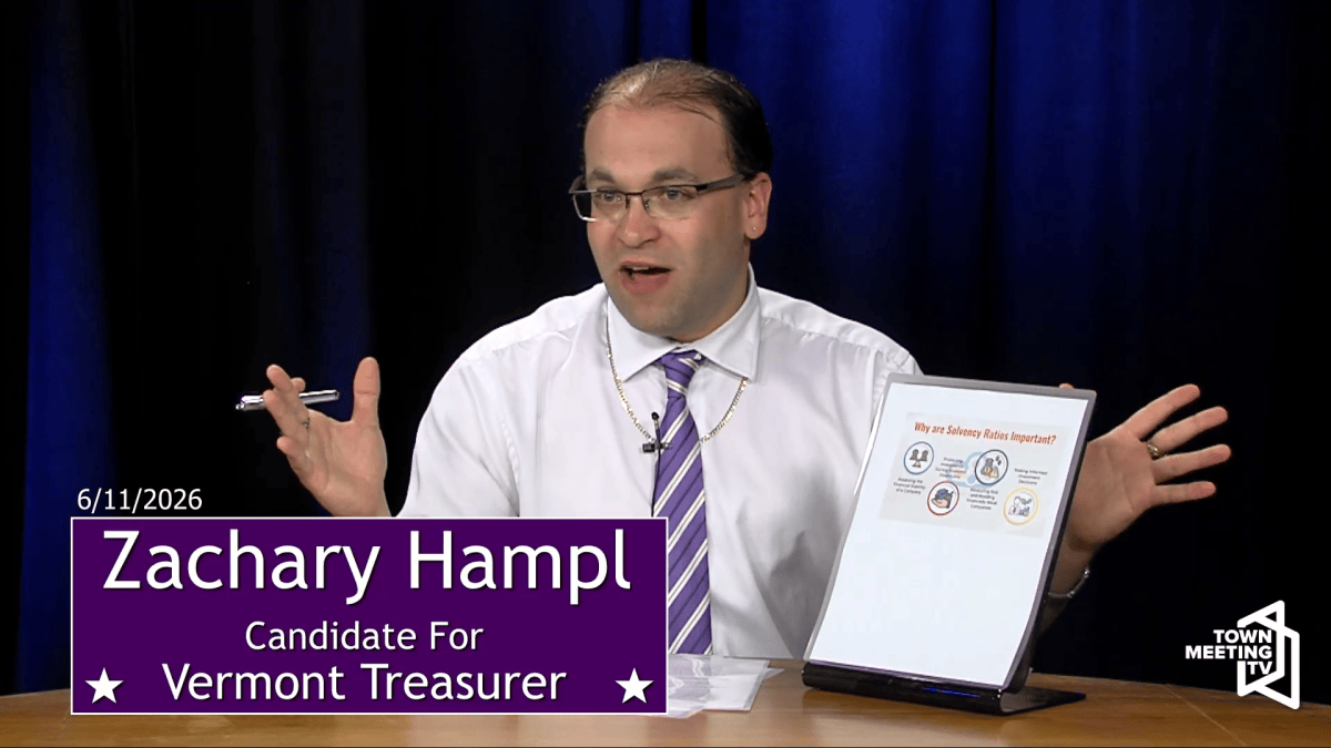 A man in a white shirt and striped tie speaks while seated at a desk with a sign reading “Zachary Hampl, Candidate for Vermont Treasurer.” He gestures toward a chart on a display.