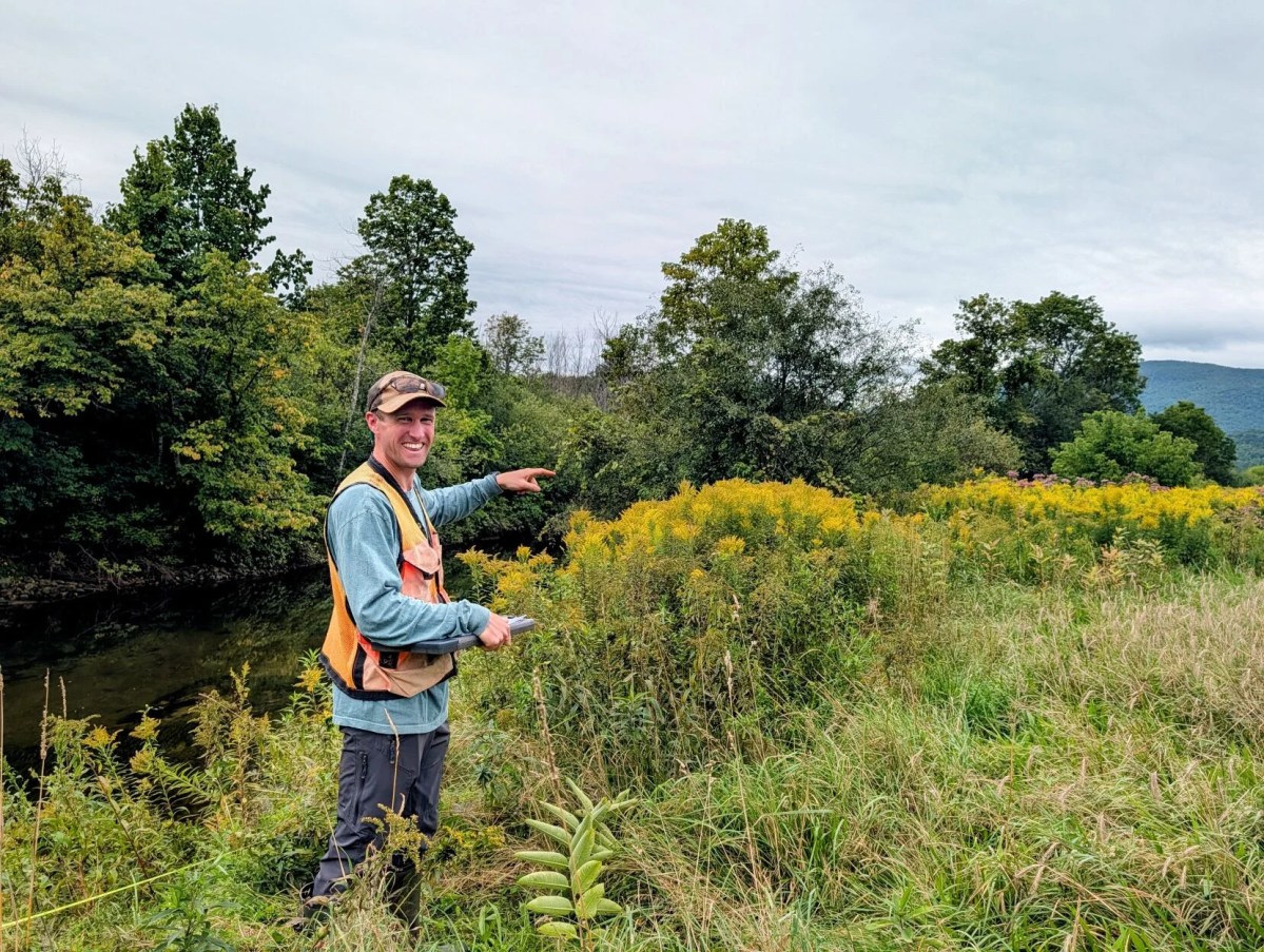 A person in outdoor gear stands in tall grass near a river, pointing toward yellow wildflowers and trees under a cloudy sky.