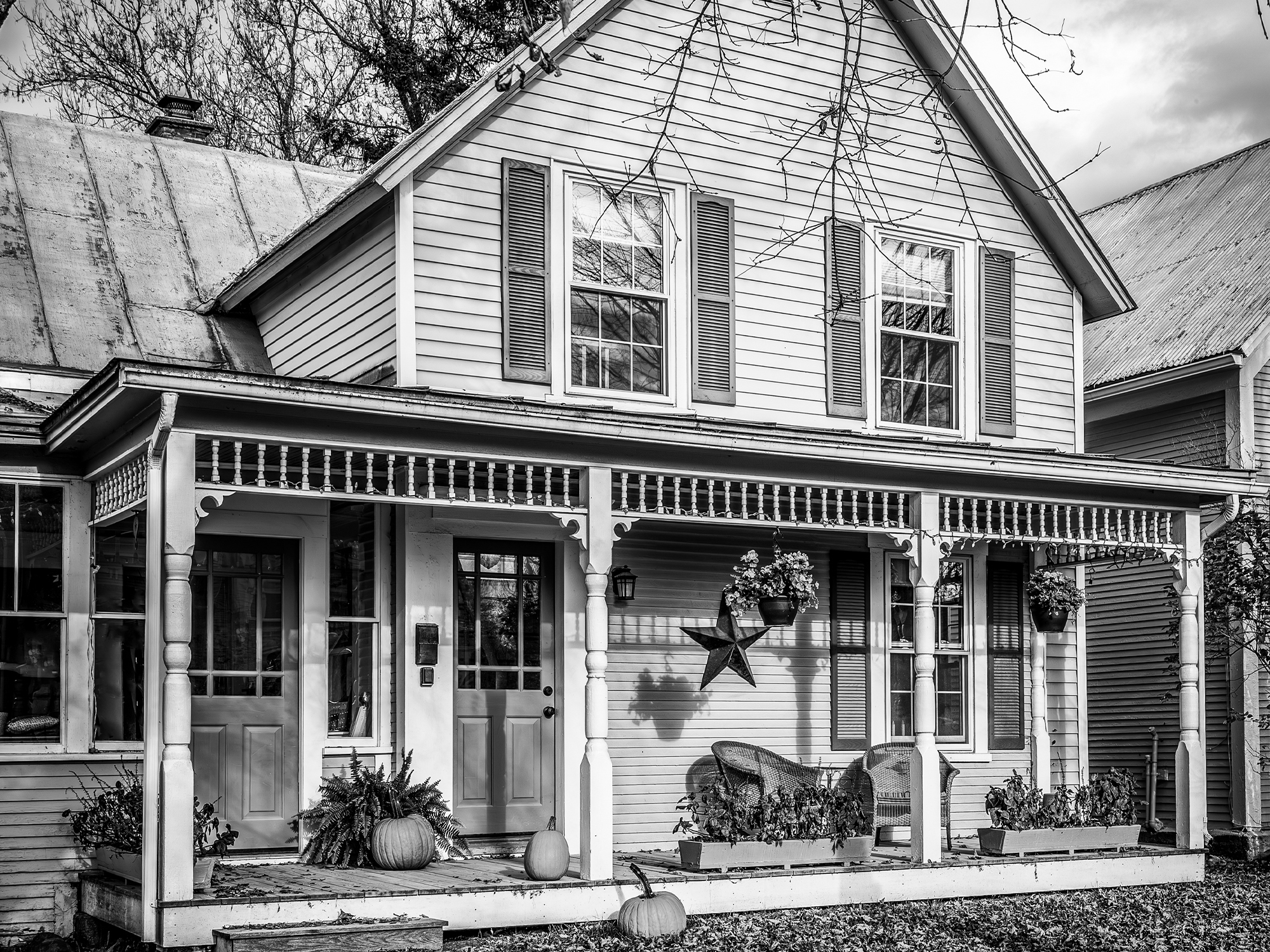 Black and white photo of a two-story house with a front porch, potted plants, hanging baskets, pumpkins, and a star decoration on the wall.