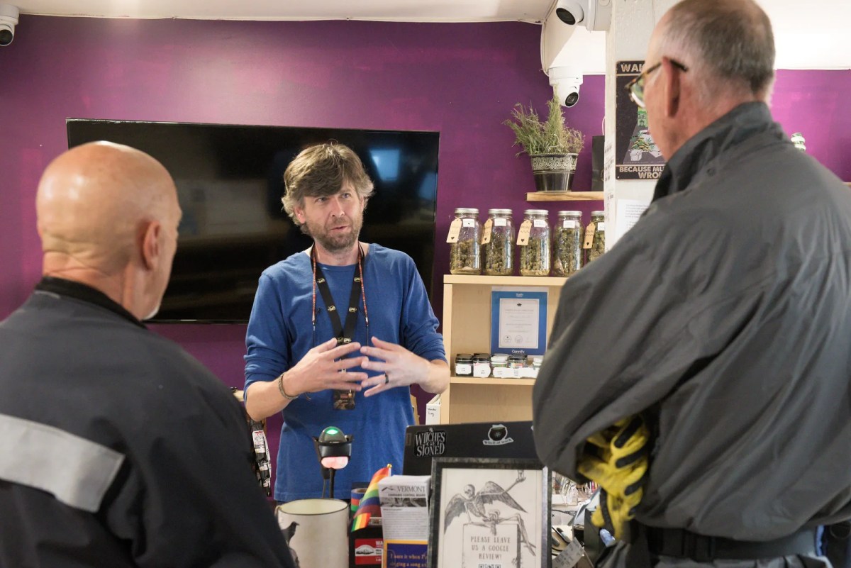 A man stands behind a counter talking to two men in jackets; shelves with jars and informational signs are visible in the background.