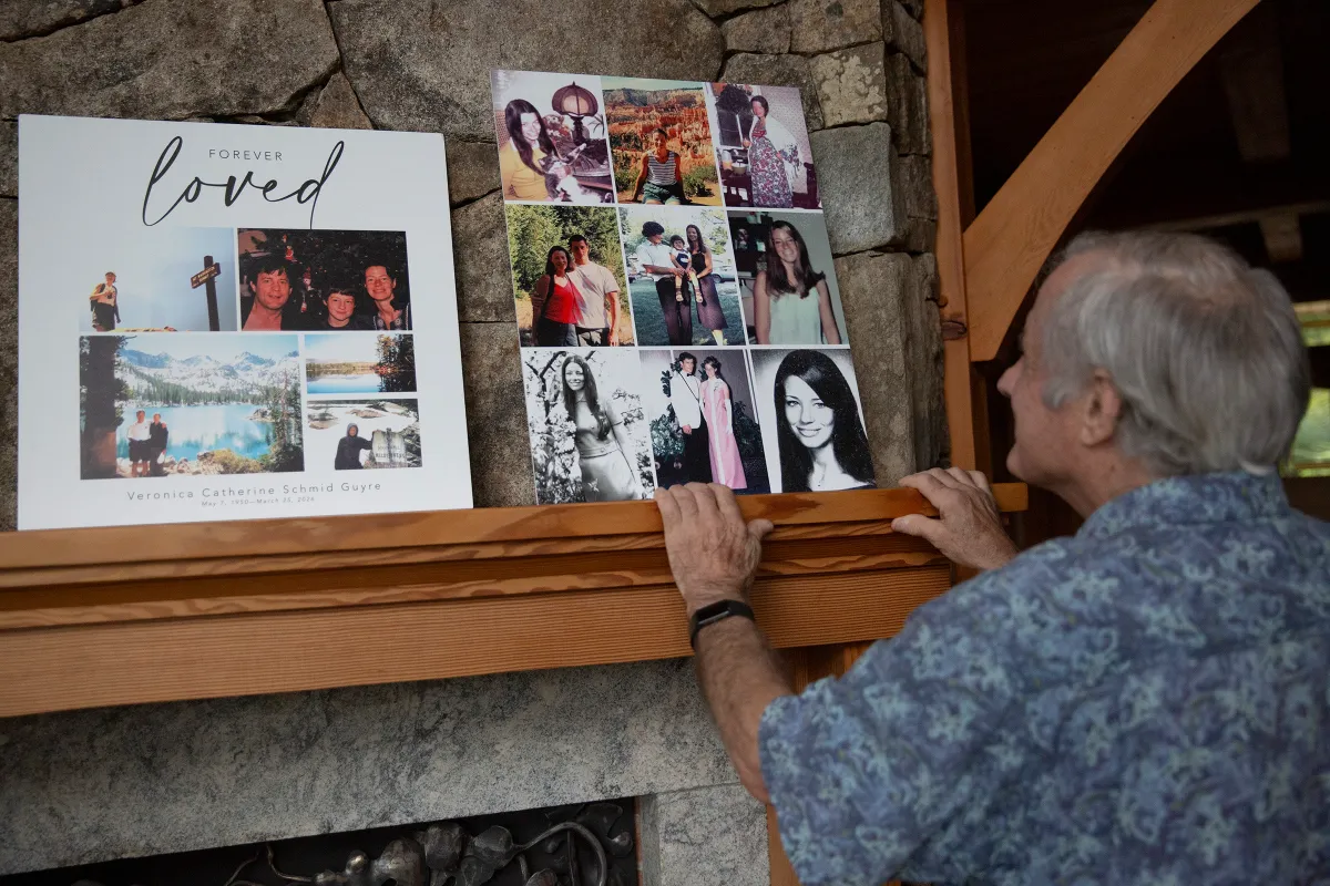 An older man looks at two memorial photo collages displayed on a mantel, one with “Forever Loved” written on it, in a home with a stone fireplace.
