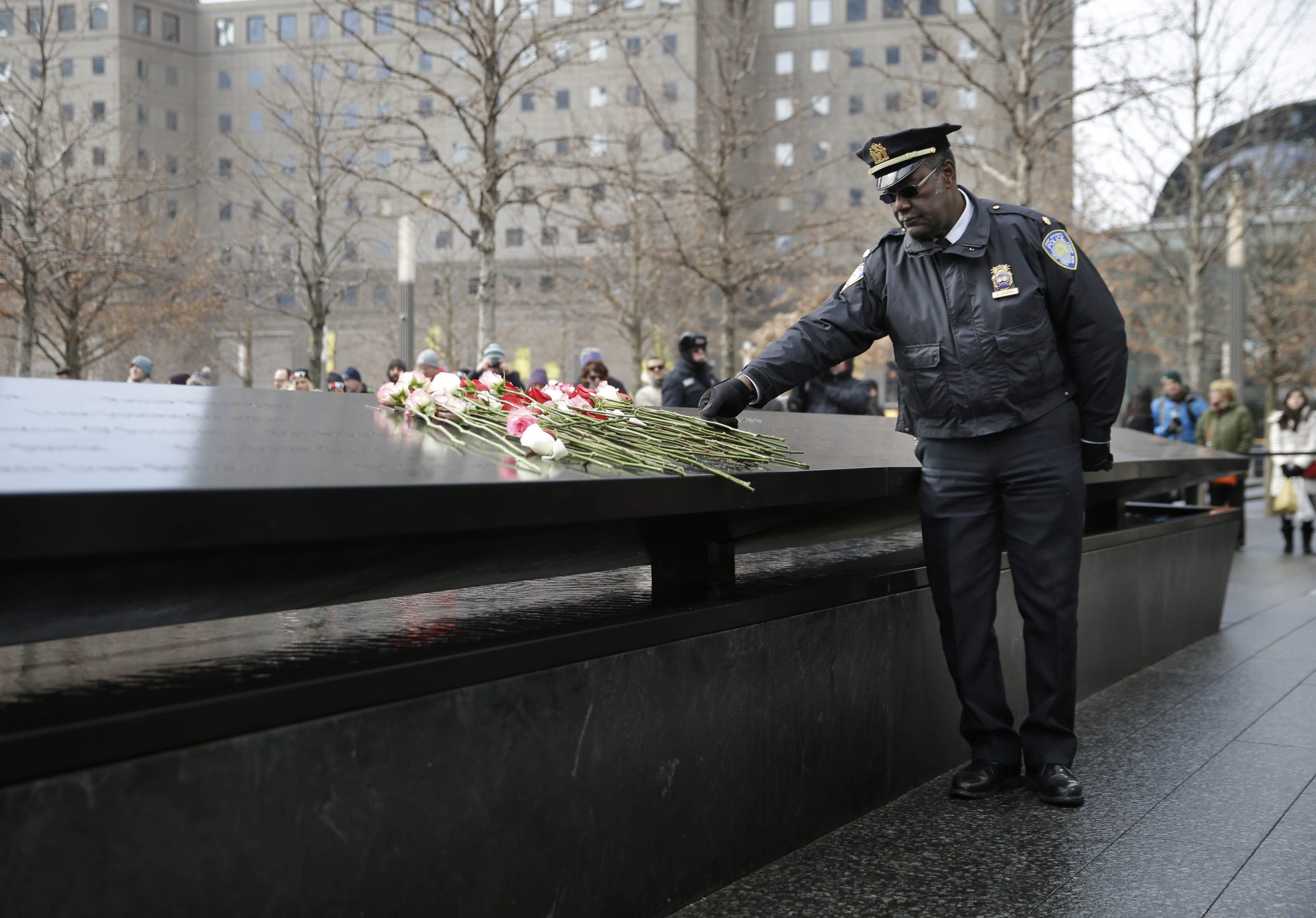 A uniformed officer places flowers on a memorial inscribed with names, with buildings and trees visible in the background.