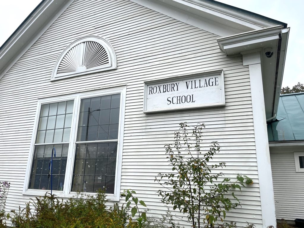 White wooden building with large windows and a sign reading “Roxbury Village School” above the entrance.
