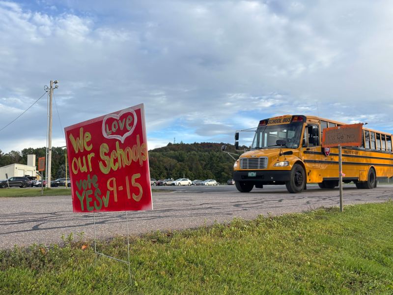 A yellow school bus drives on a road near a red sign that reads, “We love our schools. Vote YES 9-15.” The sky is partly cloudy, and grass is visible in the foreground.
