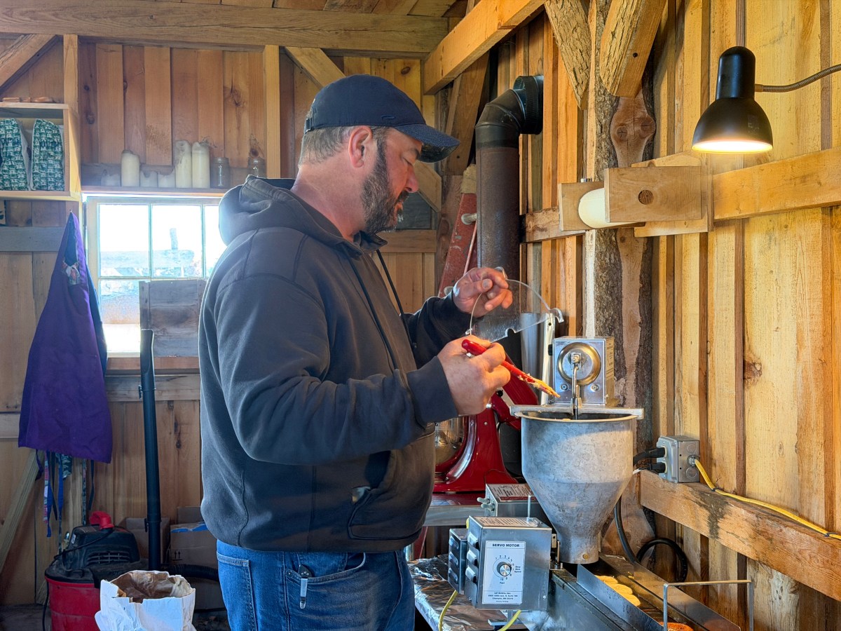 A man in a hoodie and cap operates a small metal machine inside a wooden workshop, adjusting controls under a hanging lamp.