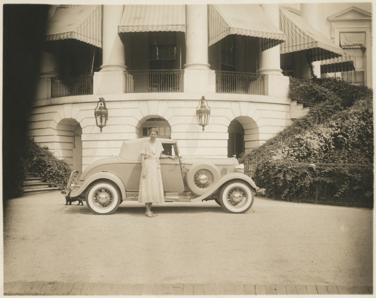 A woman in a dress stands beside a vintage convertible car parked in front of a large building with columns and striped awnings.