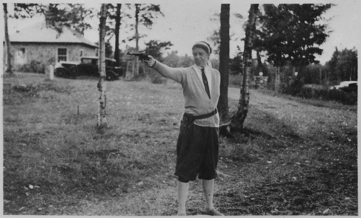 A person in vintage clothing stands outdoors on grass, aiming a handgun with one arm extended. Trees, an old car, and a building are visible in the background.