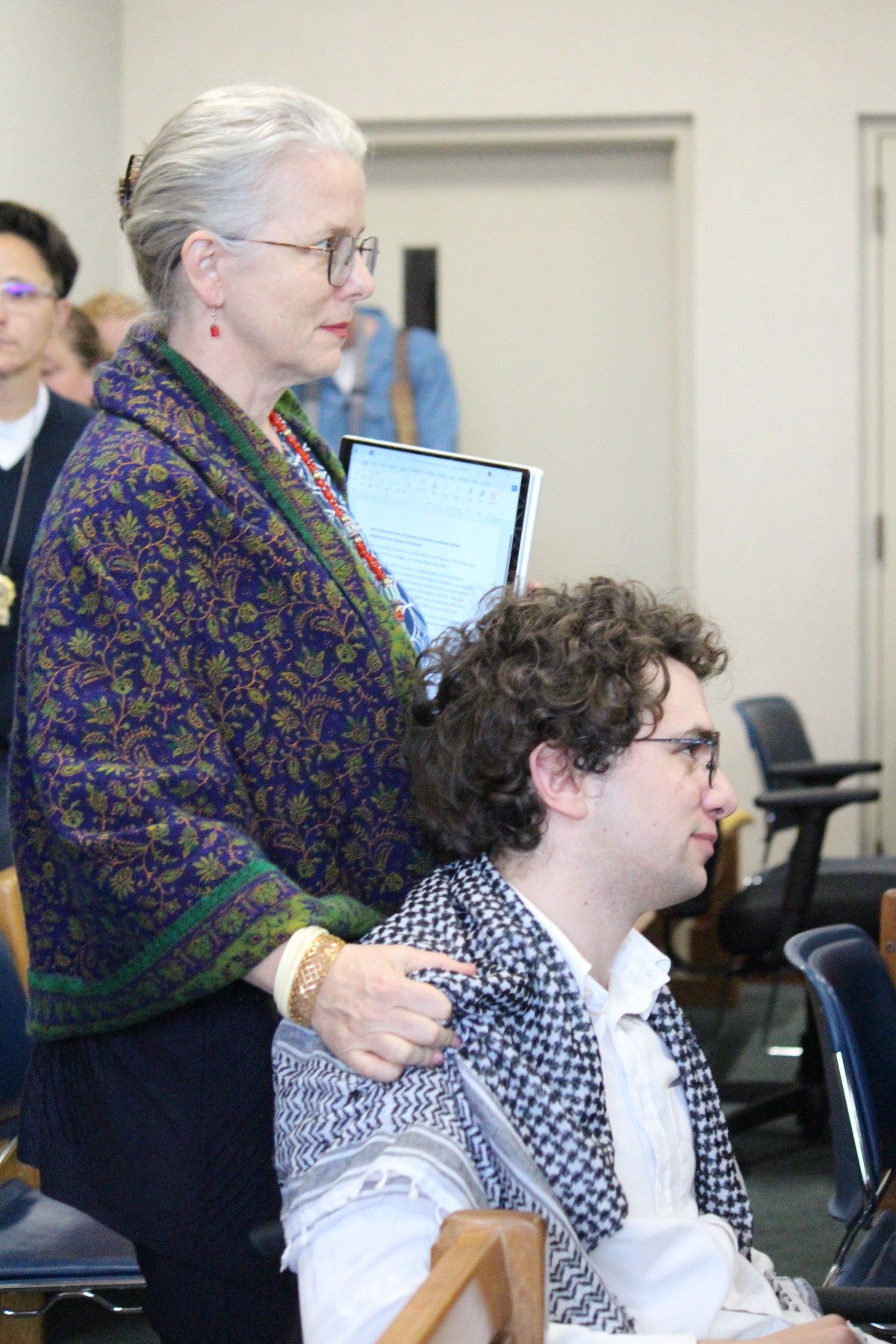 Woman stands while holding shoulder of younger man seated in front of her.
