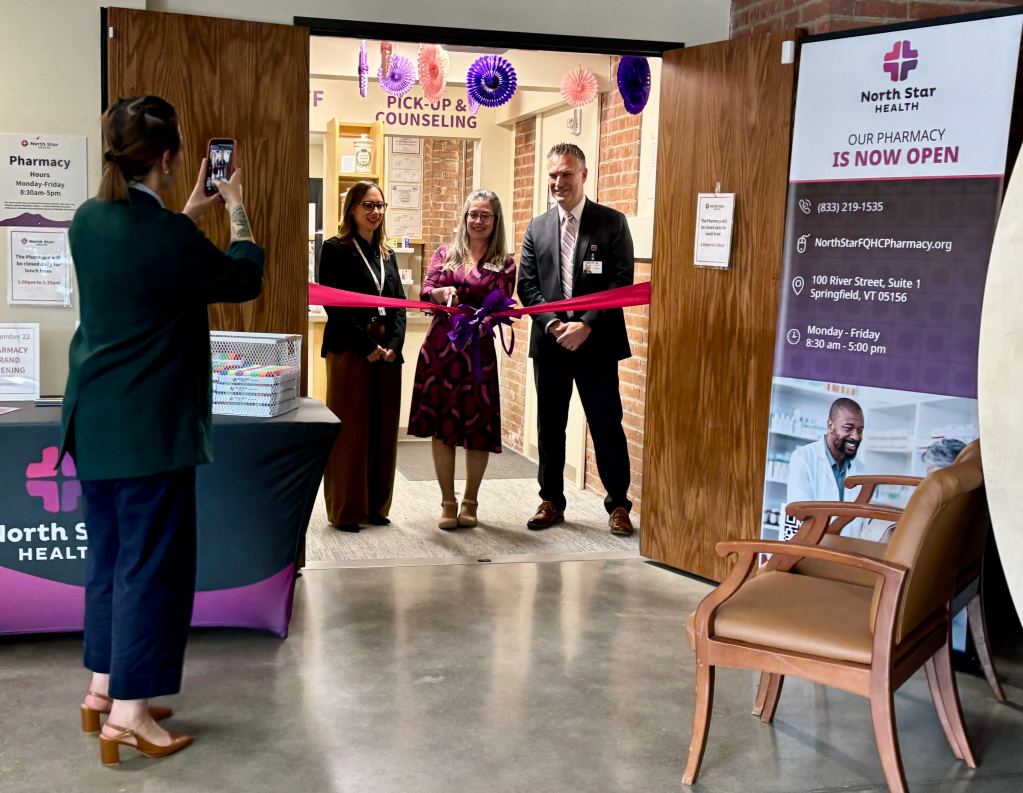 Three people stand at a ribbon-cutting ceremony in front of a pharmacy entrance while a fourth person takes their photo. Signs and balloons decorate the area.