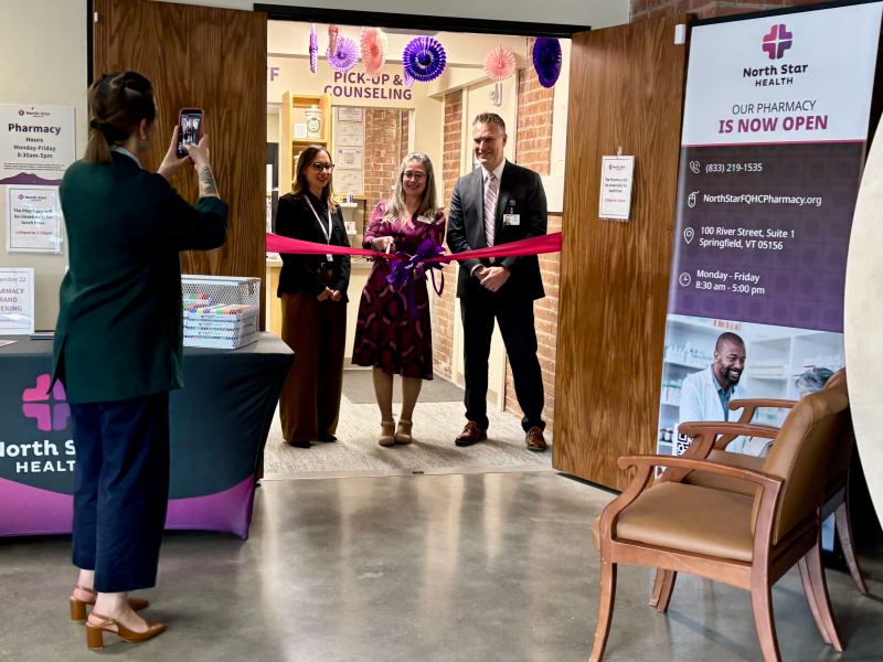 Three people stand at a ribbon-cutting ceremony in front of a pharmacy entrance while a fourth person takes their photo. Signs and balloons decorate the area.