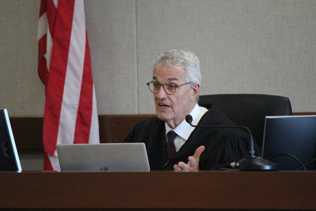 A judge in a black robe speaks while seated at a desk with laptops in a courtroom; a U.S. flag is visible in the background.