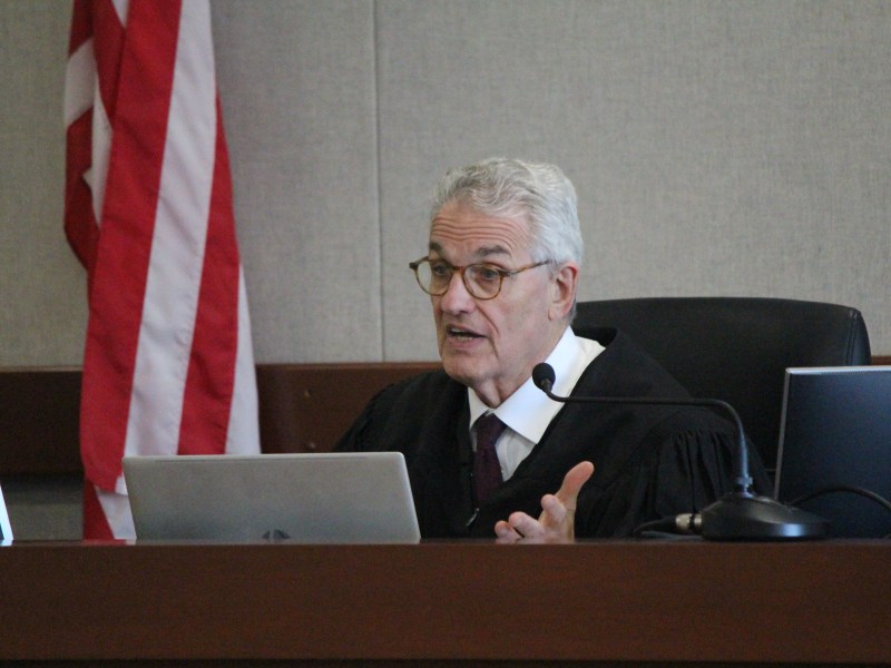 A judge in a black robe speaks while seated at a desk with laptops in a courtroom; a U.S. flag is visible in the background.