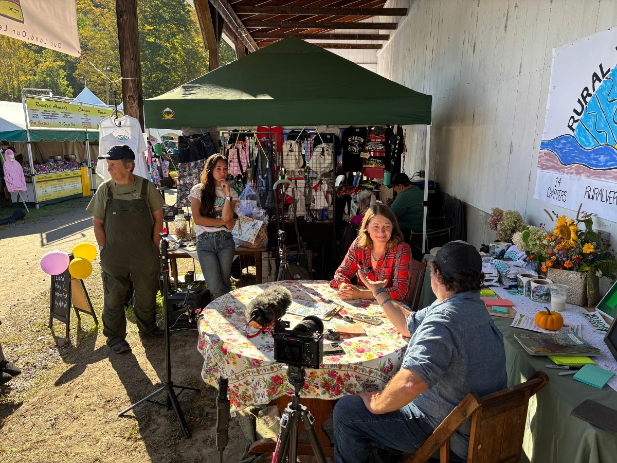 People sit at a table with recording equipment, while others stand nearby at an outdoor market stall displaying goods and informational signs.