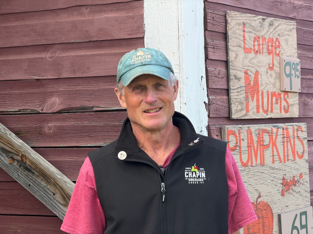 Man in a blue cap and black vest stands in front of a red wooden building with signs advertising mums and pumpkins.