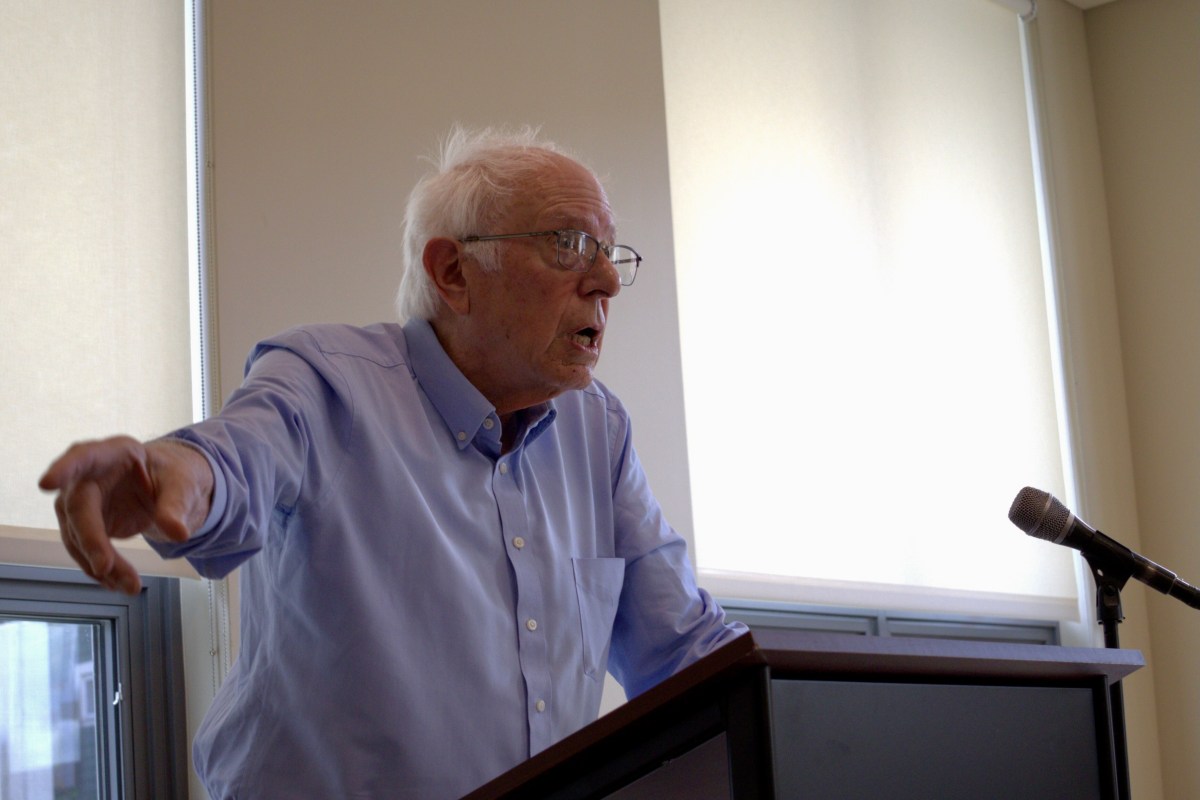 An older man with white hair and glasses speaks at a podium next to a microphone in a room with large windows and beige shades.