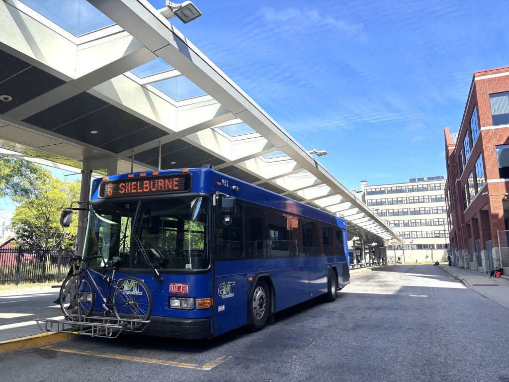 A blue city bus labeled “Shelburne” is parked at a bus terminal, with two bicycles mounted on its front bike rack under a glass canopy.