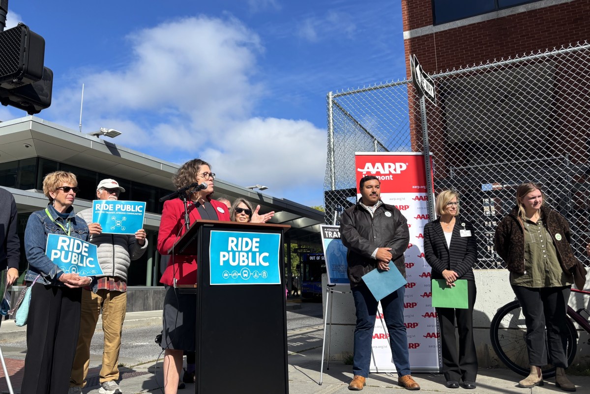 A group of people stand outdoors at a podium with a “Ride Public” sign, giving a speech near an AARP banner and transit station.