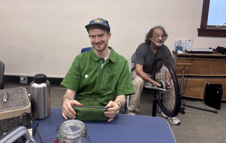 Two men are indoors; one sits at a table smiling, holding a pouch, while the other sits behind him, repairing a bicycle wheel.