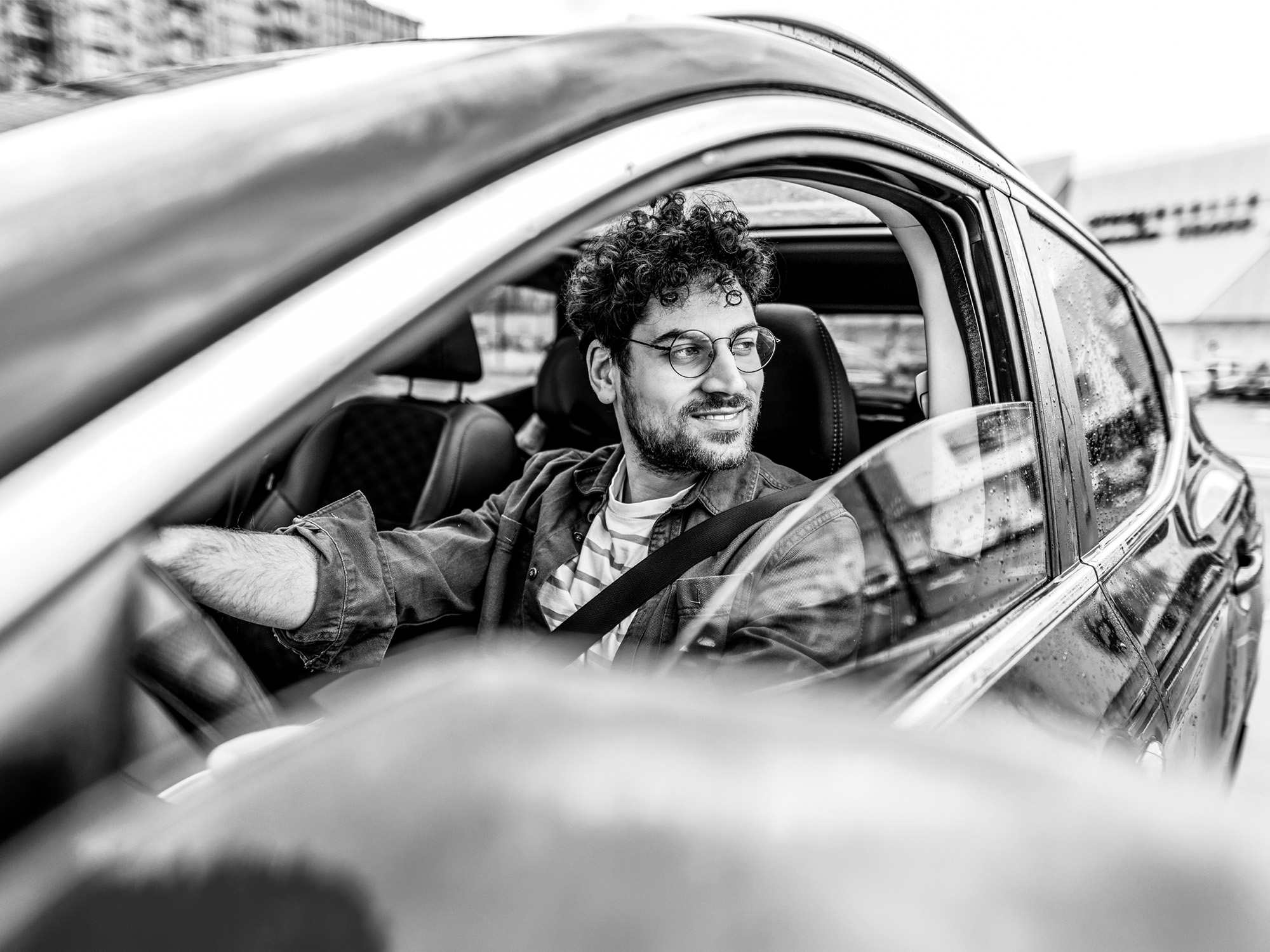A man wearing glasses sits in the driver’s seat of a car, looking out the open window and smiling. The photo is in black and white.