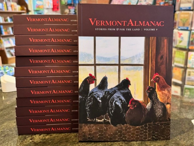 A stack of Vermont Almanac books is displayed, with the cover featuring chickens gathered by a window overlooking a rural landscape.