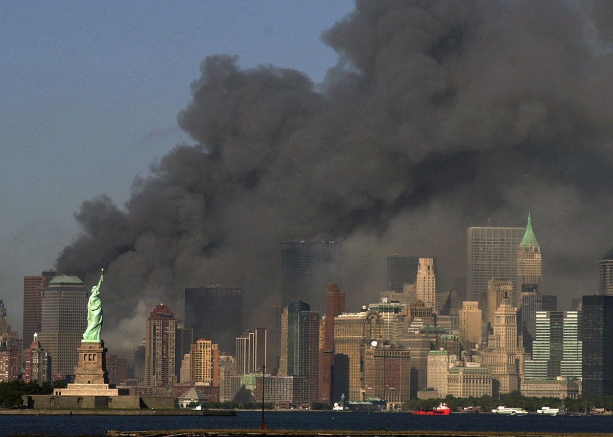 Dense black smoke rises from buildings in lower Manhattan behind the Statue of Liberty against a clear blue sky.
