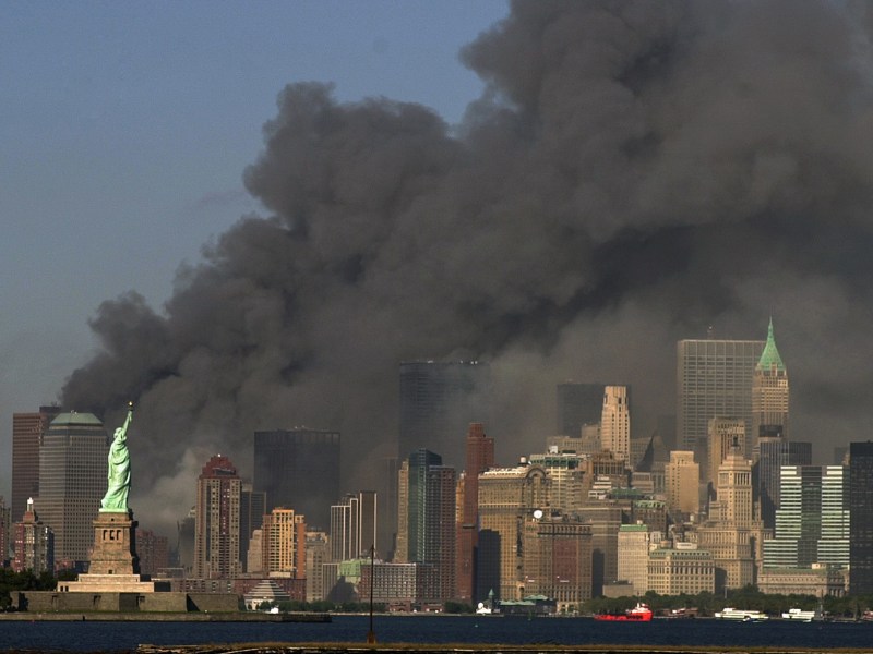 Dense black smoke rises from buildings in lower Manhattan behind the Statue of Liberty against a clear blue sky.