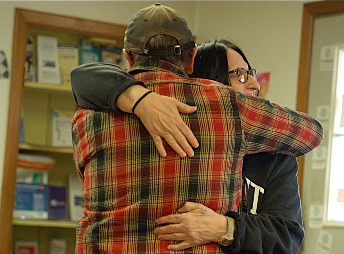 Two people, one wearing a plaid shirt and cap, and the other in glasses, are embracing in a room with shelves and posters in the background.
