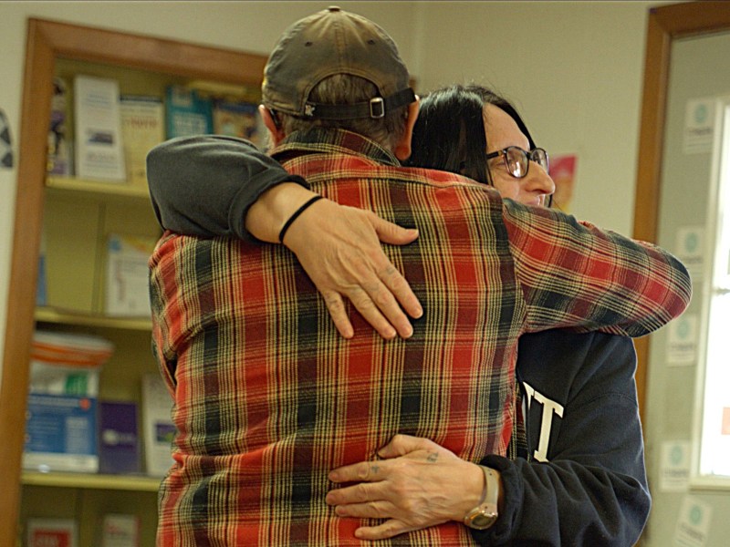 Two people, one wearing a plaid shirt and cap, and the other in glasses, are embracing in a room with shelves and posters in the background.